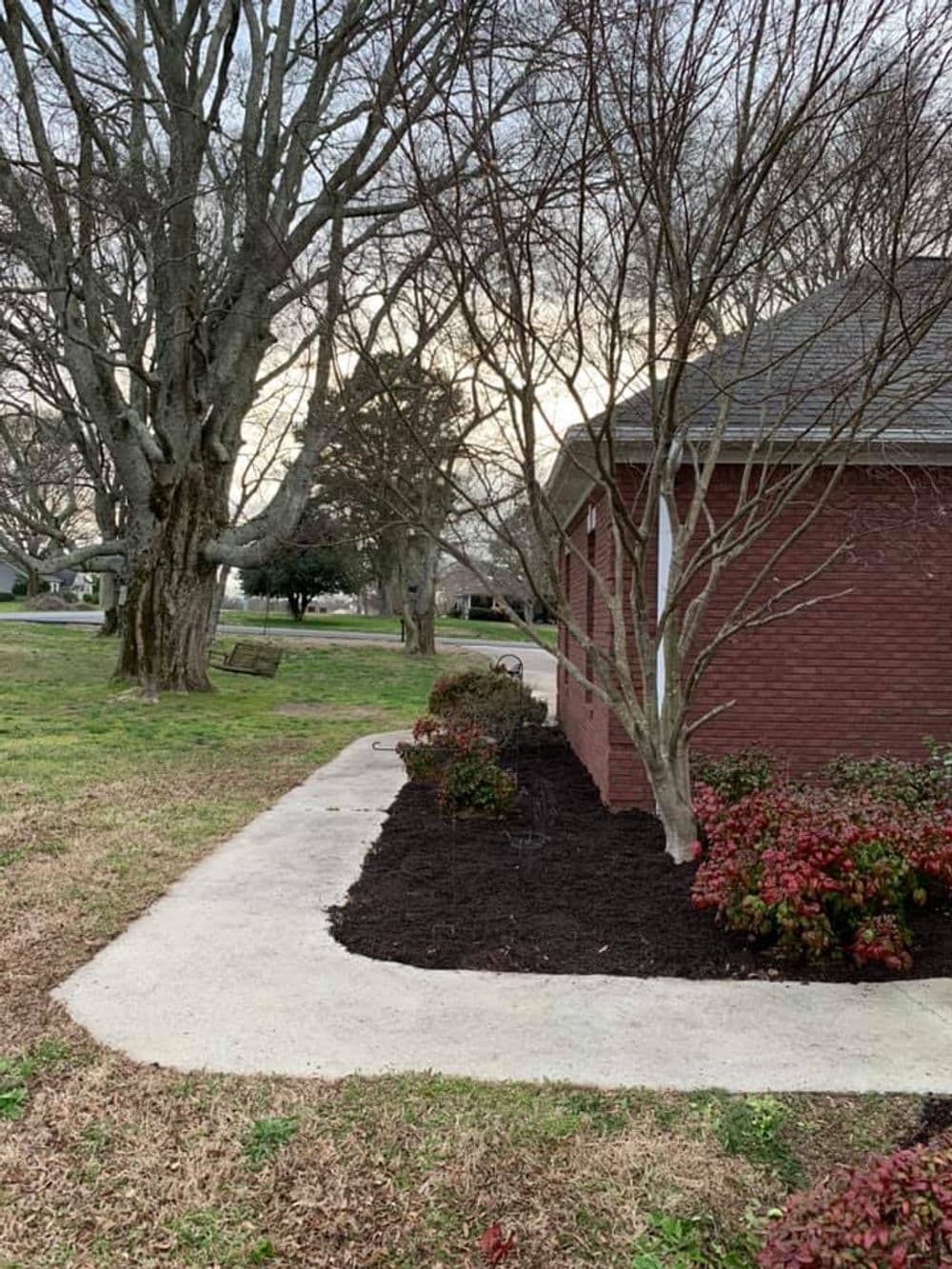 Freshly mulched flower bed alongside a brick house with surrounding trees and lawn.