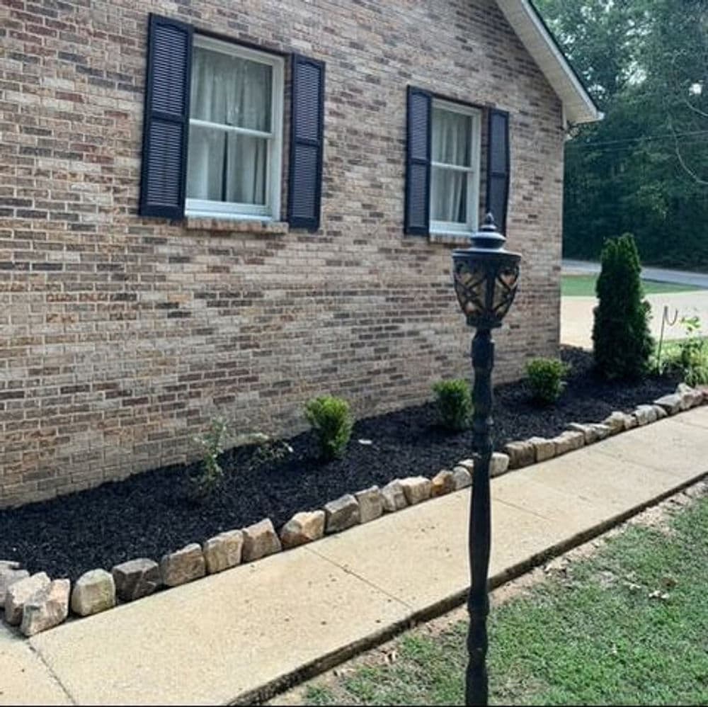 Landscape featuring brick house, green shrubs, and stone-bordered flowerbed with lantern.