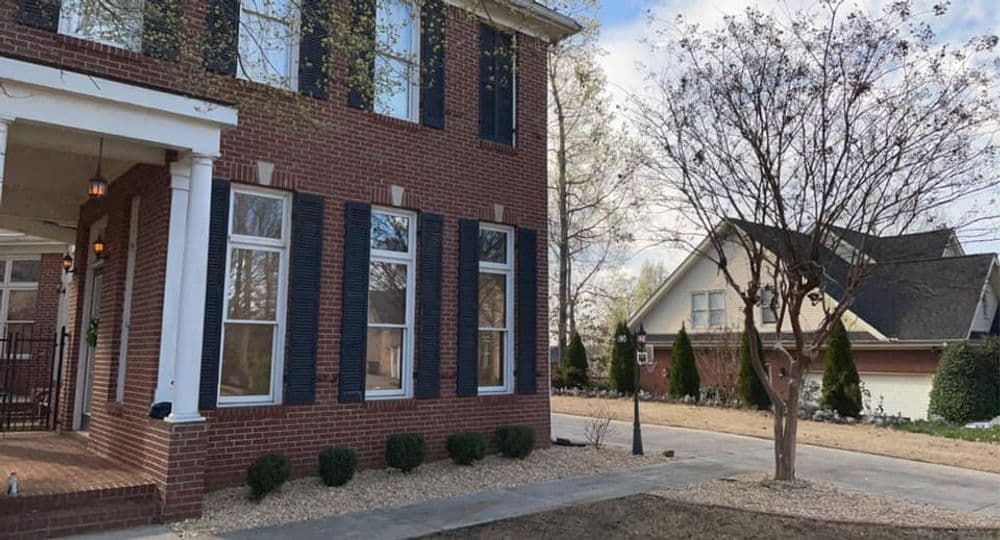 Brick house with black shutters and landscaped yard, featuring trees and neighboring homes.