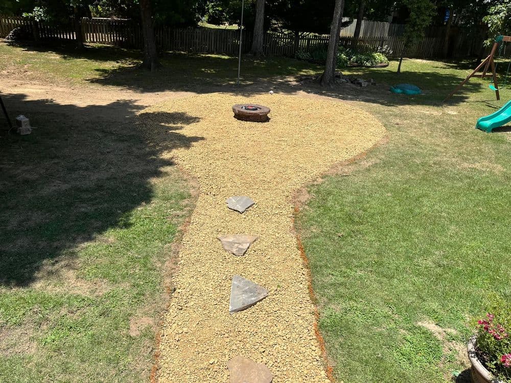 Gravel pathway with stone steps leading to a fire pit in a grassy backyard setting.