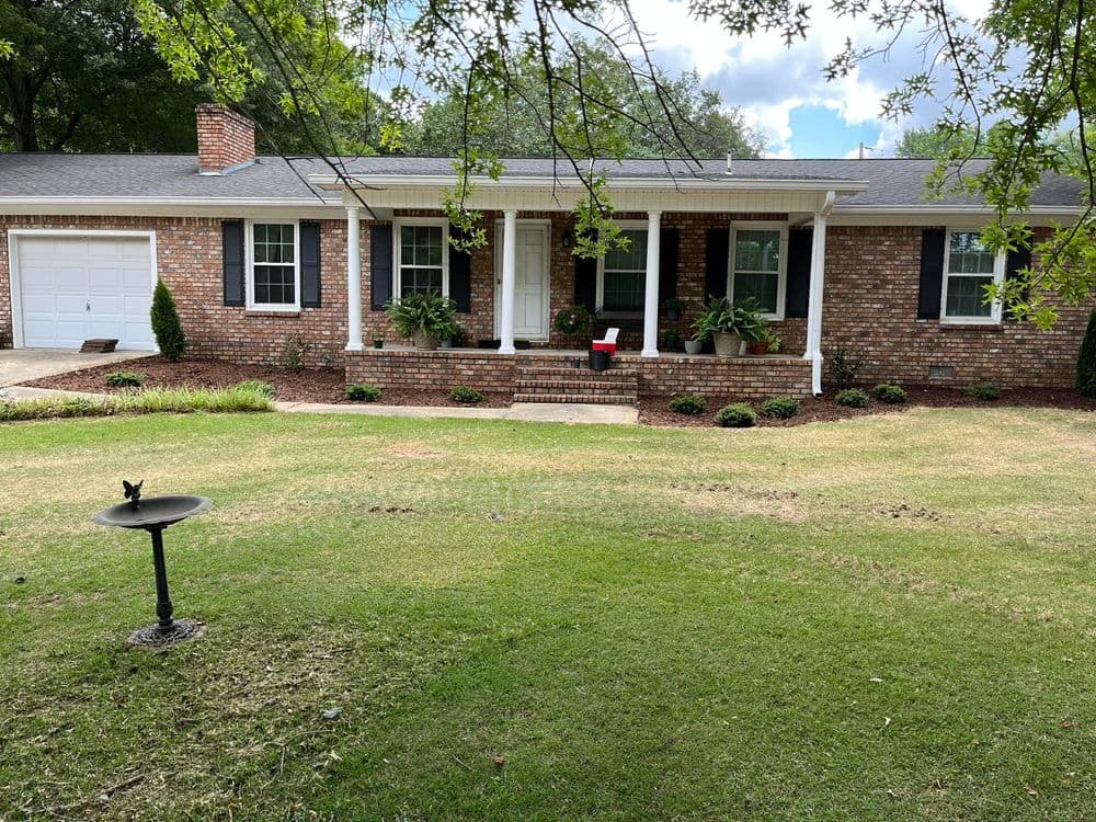 Brick house with white porch, green lawn, and driveway, surrounded by trees and shrubs.