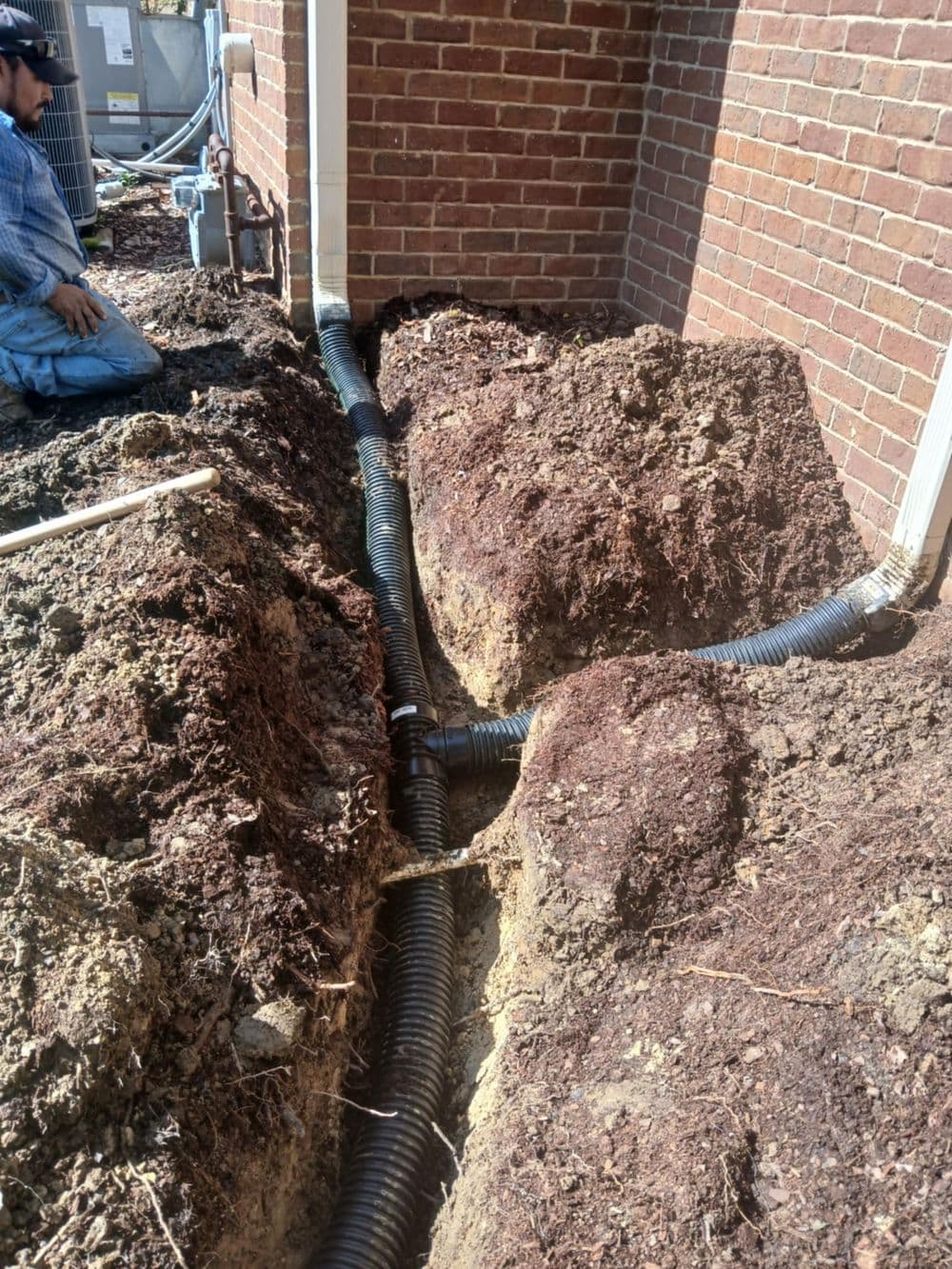 Drainage system installation with trenches and PVC piping near a brick foundation.
