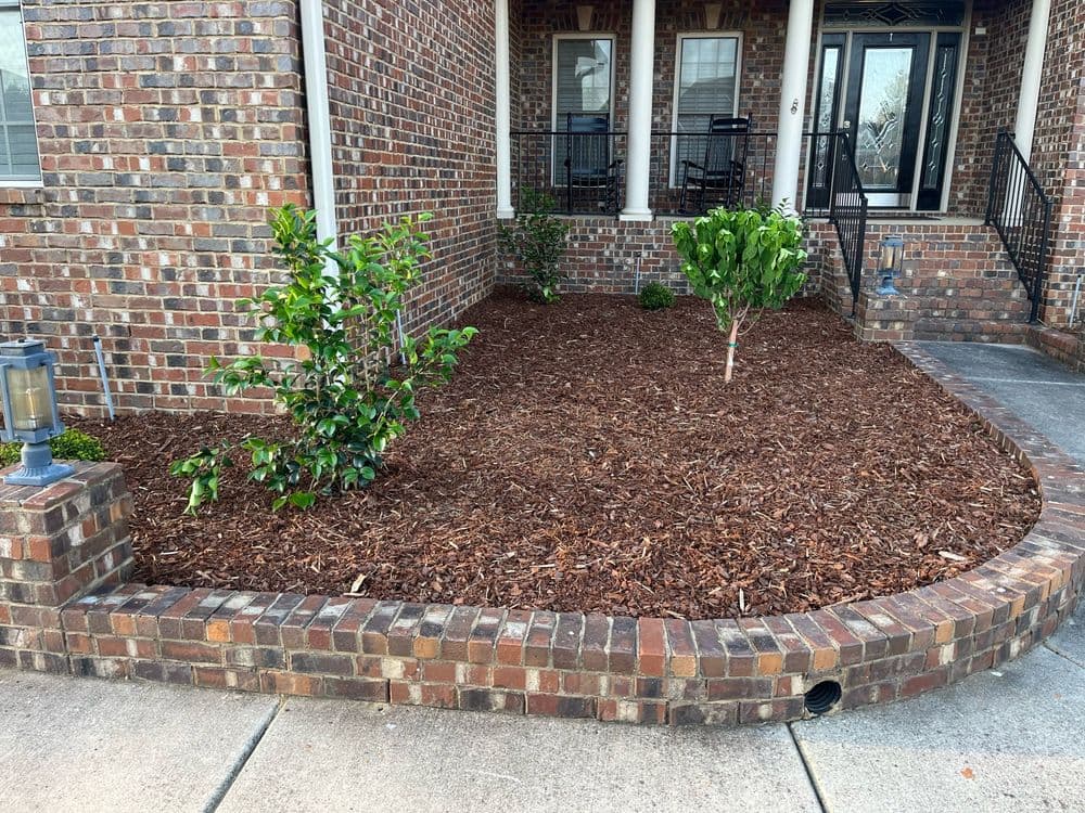 Well-maintained front yard with mulch, shrubs, and a brick border of a brick house.