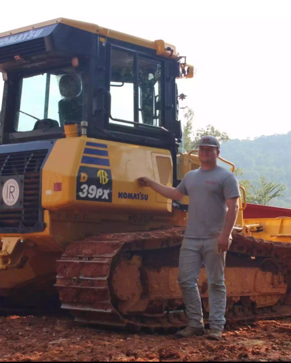 Man standing beside a Komatsu D39PX bulldozer on a construction site in a wooded area.