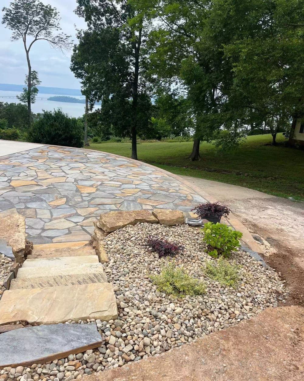 Stone pathway leading to a landscaped garden with rocks and greenery near a lake.