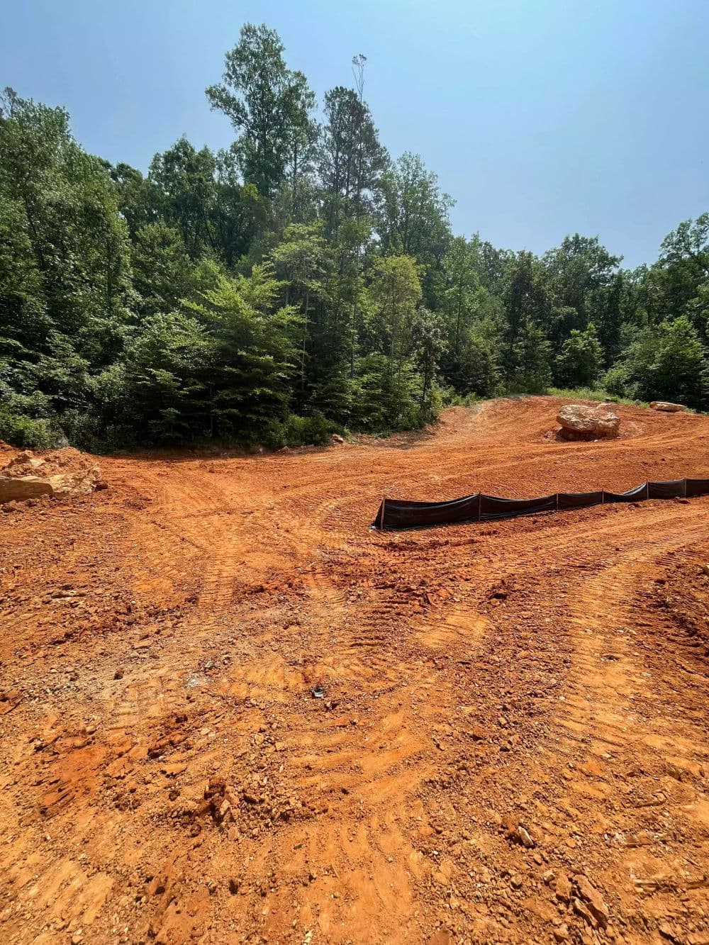 Construction site with red dirt, tree line, and silt fence in wooded area under clear sky.